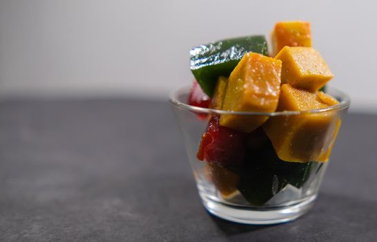 Close-up of glass of red, yellow, and green fruit paste slices above black and white background. Authentic natural Mexican candy cubes in cup above dark surface. Tasty traditional snacks