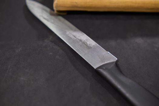 Close-up of long knife with black plastic handle on dark surface. Sharp kitchen utensil with slightly worn blade isolated. Kitchenware and food preparation
