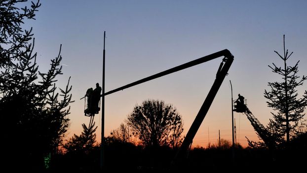 Silhouettes of male workers installing a street lamp on a crane machine at sunset to illuminate the road and street. Electricians install street lighting lines