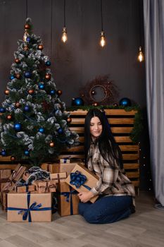 confident beautiful young woman in checkered shirt and jeans near christmas tree decorated in blue tones.