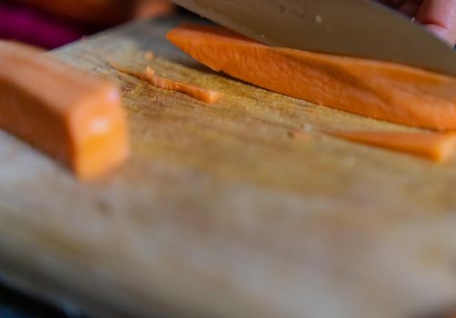 Close-up of hands cutting yam slices on wooden cutting board. Person slicing fresh orange sweet potato above wooden surface. Healthy meal preparation