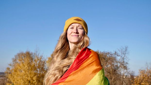 Portrait of a young, Caucasian, blonde, girl with acne holding a rainbow LGBT flag in autumn. Woman in yellow looking at camera on a sunny day