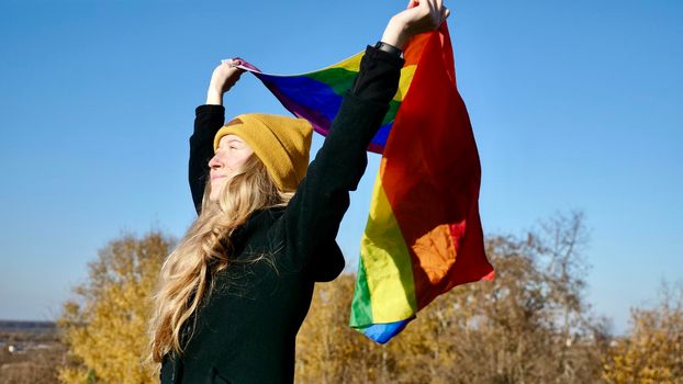 Portrait of a young, Caucasian, blonde, girl with acne holding a rainbow LGBT flag in autumn. Woman in yellow hat and black coat on a sunny day