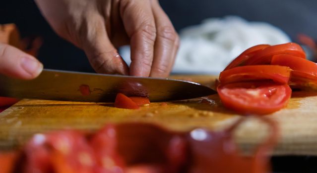 Close-up of hands firmly slicing tomato on wooden cutting board with blurry sliced onion as background. Person cutting fresh red vegetable with big knife above wooden surface. Healthy meal preparation