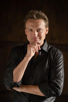 Confident Wealthy Man in Black Leather Shirt Posing for the Camera in the Studio on Brown Background. Male Model of 35s with Blond Hair and Blue Eyes Looks at the Camera. Close-up Portrait. High quality photo