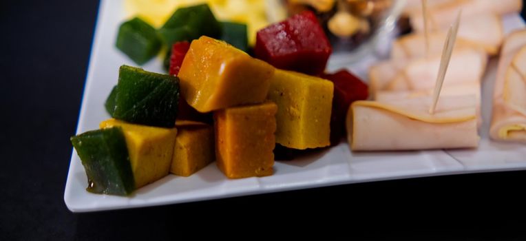 Wide view of diced fruit paste, turkey ham rolls, and cheddar cheese cubes on square white plate. Close-up of turkey meat, colorful Mexican candy, and sliced cheese above black surface. Healthy snacks