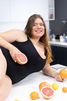 Female Model Plus Size in Black Swimsuit Posing on the Kitchen Table with Citrus Fruits Orange, Grapefruit, Mandarin. Healthy Food, Body Positive. Close-up. High quality photo