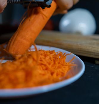 Close-up of hands peeling carrot with julienne peeler above white plate with blurry vegetables as background. Person making small pile of grated orange vegetables. Healthy food preparation
