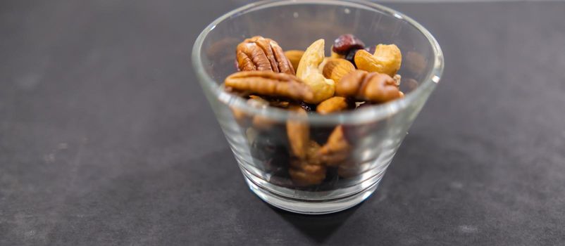 Wide view of small glass of walnuts, almonds, Indian nuts, and dry cranberries on black surface. Close-up of fresh natural nuts in glass cup above dark background. Tasty traditional snacks