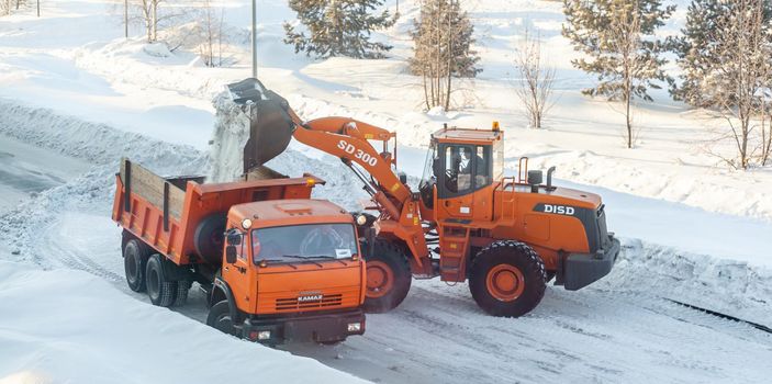 December 23, 2021. Kemerovo Region, Russia. Big orange tractor cleans up snow from the road and loads it into the truck. Cleaning and cleaning of roads in the city from snow in winter. 