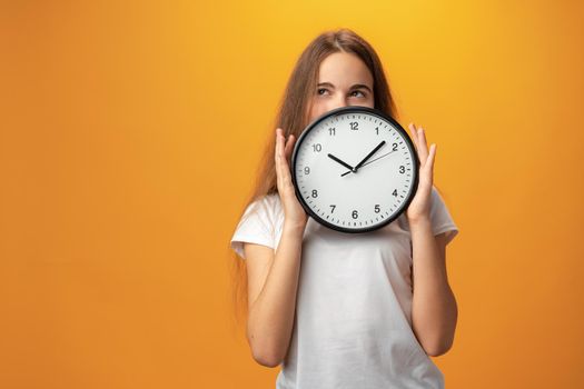 Beautiful teen girl holding wall clock over yellow background, close up