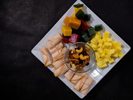Top view of turkey ham rolls, diced fruit paste, cheddar cheese cubes, and glass of walnuts on square white plate. Turkey meat, colorful Mexican candy, and nuts above black surface. Healthy snacks