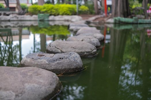 Close-up of stepping stones path on pond with blurry trees as background. Peaceful view of greenish water and rocks in Masayoshi Ohira Park. Classic architecture and outdoors