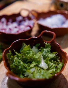 Close-up of lettuce and blurry chopped onions, and radishes in clay pots on table. Fresh sliced vegetables in handmade clay bowls on wooden surface. Traditional Mexican cuisine