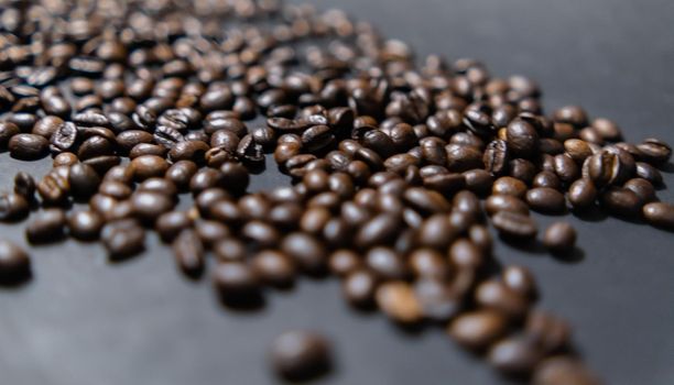 Close-up of small pile of roasted coffee beans on dark gray surface. Group of coffee grains scattered above dark color background. Caffeine and drink preparation