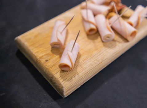 Close-up of turkey ham rolls with toothpicks and walnuts on cutting board. Wooden board with fresh turkey breast meat and nuts above black surface. Healthy meal preparation