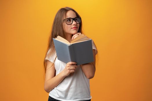 Smart teen girl in glasses holding book against yellow background, close up