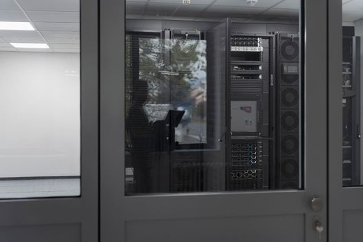 Software Engineer Working on a Laptop Computer in a Modern Server room. Monitoring Room Big Data Scientist in reflection of the entrance door. High quality photo