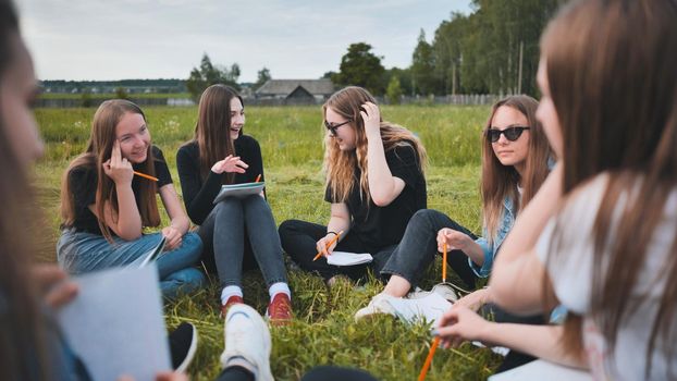 A group of female students are sitting in a circle on a meadow for collective work with notebooks