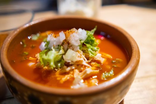 Close-up of clay bowl of delicious and traditional red pozole on wooden table. Authentic Hispanic pork stew in handmade bowl with chopped onion and lettuce on top. Traditional Mexican cuisine