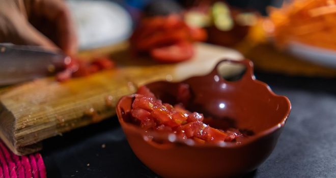 Close-up of chopped tomato in small clay pot with hands slicing more tomato as background. Person cutting fresh red vegetable with big knife on wooden cutting board. Healthy meal preparation