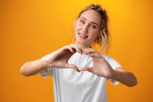 Young beautiful woman showing heart symbol over yellow background, close up