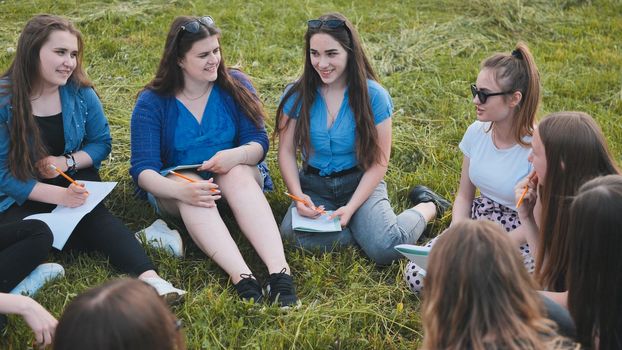 A group of female students are sitting in a circle on a meadow for collective work with notebooks