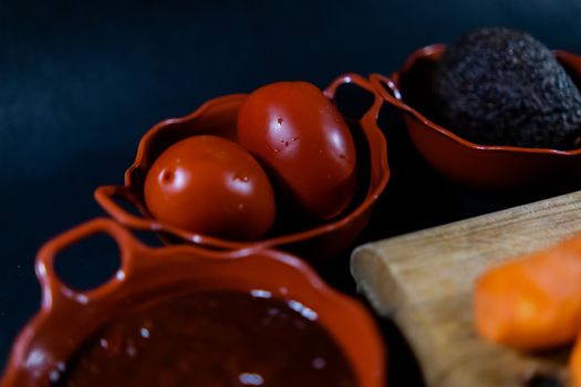 Close-up of hot sauce, two tomatoes, and avocado in traditional clay pots with black background. Fresh vegetables and spicy condiments above dark surface. Authentic food preparation
