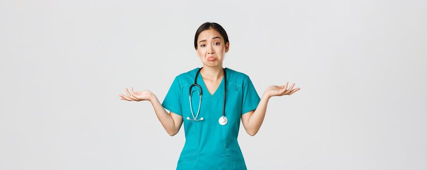 Covid-19, healthcare workers, pandemic concept. Portrait of clueless asian female nurse, woman doctor shrugging and spread hands sideways unaware, dont know, cant help, white background.