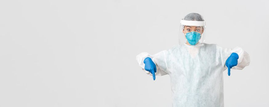 Covid-19, coronavirus disease, healthcare workers concept. Surprised and excited female doctor, physician in personal protective equipment pointing fingers down, standing white background.