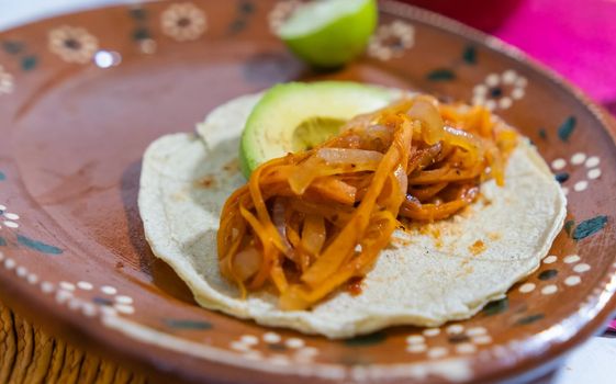 Close-up of halved lime, avocado slice, and vegan tinga taco on handmade clay plate. Authentic spicy sliced carrots and onion dish on brown plate above colorful table mats. Classic Mexican cuisine