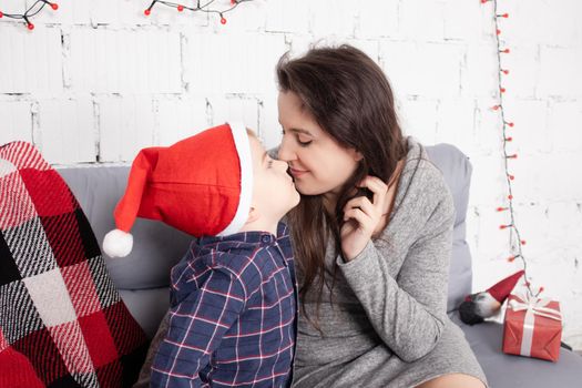 happy incomplete family - mother and son - hugging on grey couch covered in red checkered plaid-blanket near red christmas decor. cozy young family. single mother.