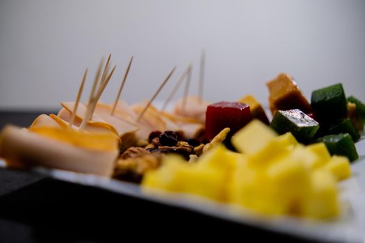Close-up of turkey ham rolls, diced fruit paste, cheddar cheese cubes, and walnuts on square white plate. Turkey meat, colorful Mexican candy, and nuts with white background. Healthy snacks