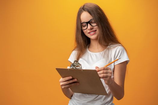 Teen school girl taking notes on clipboard against yellow background, close up