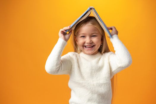 Little girl holding book on her head and looking at camera over yellow background, close up