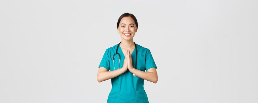 Covid-19, healthcare workers and preventing virus concept. Smiling beautiful asian female nurse, physician in scrubs smiling, hold hands together over chest in namaste, greeting gesture.