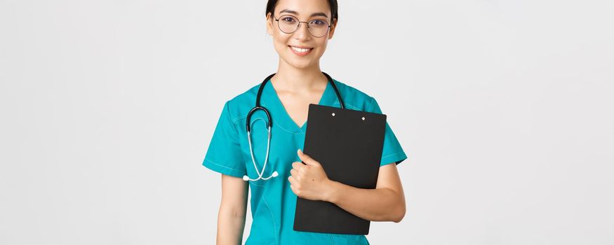 Covid-19, coronavirus disease, healthcare workers concept. Smiling confident asian female nurse, physician in glasses and scrubs holding clipboard, making examination, white background.