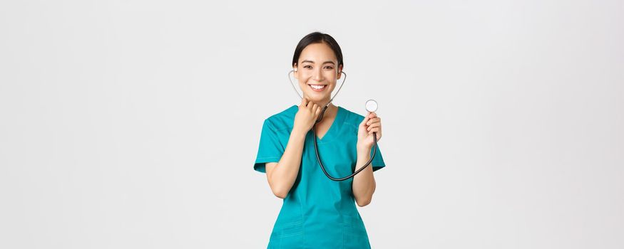 Covid-19, healthcare workers and preventing virus concept. Cheerful smiling asian female physician, doctor consult patient, examine person with stethoscope, standing white background.