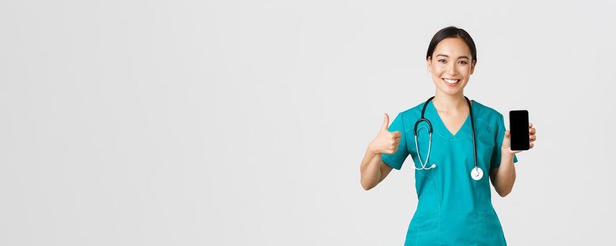 Covid-19, healthcare workers and online medicine concept. Portrait of young asian female physician, doctor in scrubs show thumbs-up and smartphone screen, promote application, white background.