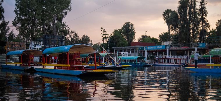 Astonishing view of traditional colorful trajineras in Xochimilco lake under beautiful cloudy sky. Authentic roofed boats with wooden chairs and tables in canal. Mexican traditions and culture