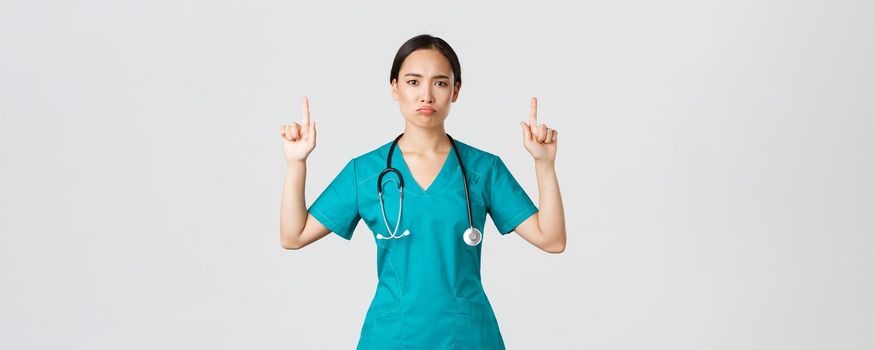 Covid-19, healthcare workers, pandemic concept. Gloomy disappointed asian female doctor, physician in scrubs pointing fingers up and grimacing displeased, standing white background.