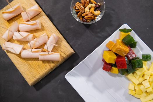 Top view of turkey ham rolls, diced fruit paste, cheese cubes, and walnuts above black surface. Fresh turkey meat, colorful diced food, and nuts on wooden boards, plates, and glass cups. Balanced diet
