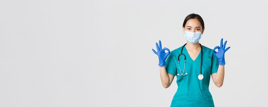 Covid-19, coronavirus disease, healthcare workers concept. Smiling professional and confident asian doctor, physician in medical mask, gloves and scrubs show okay gesture, guarantee quality service.