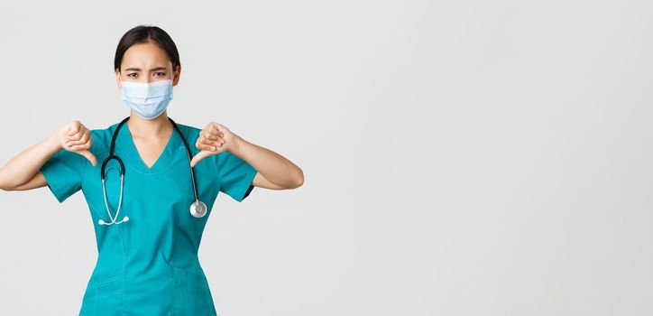 Covid-19, coronavirus disease, healthcare workers concept. Disappointed and upset asian female doctor in scrubs and medical mask showing thumbs-up, disagree or prohibit something, give advice.