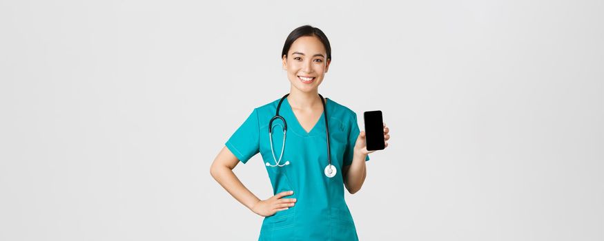 Covid-19, healthcare workers and online medicine concept. Professional female asian doctor, nurse in scrubs showing smartphone screen and smiling, showing internet appointment app, white background.