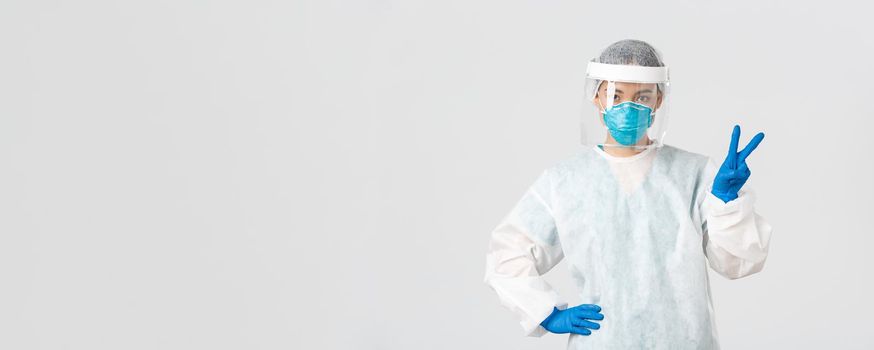 Covid-19, coronavirus disease, healthcare workers concept. Sassy asian female doctor, physician or tech lab in personal protective equipment showing peace sign, standing white background.