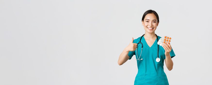 Healthcare workers, preventing virus, quarantine campaign concept. Confident smiling asian female nurse, doctor in scrubs recommend medication, show thumbs-up and pills, white background.