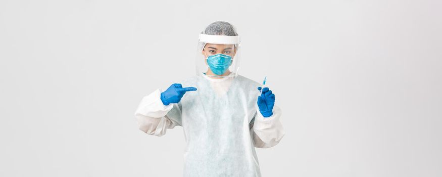 Covid-19, coronavirus disease, healthcare workers concept. Serious asian female doctor, physician in PPE personal protective equipment, pointing finger at syringe with vaccine, white background.