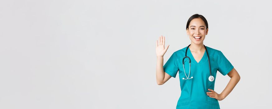 Covid-19, healthcare workers and preventing virus concept. Friendly-looking smiling asian female doctor, physician in scrubs waving hand to say hi, hello, greeting patient, nice to see you.