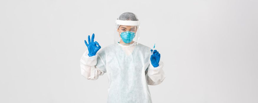 Covid-19, coronavirus disease, healthcare workers concept. Confident serious female asian doctor in personal protective equipment, show okay gesture, hold syringe with vaccine, white background.
